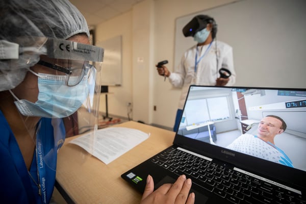 Nursing student in mask and face shield controls UbiSim virtual reality scenario while other student in mask stands with headset and hand controllers